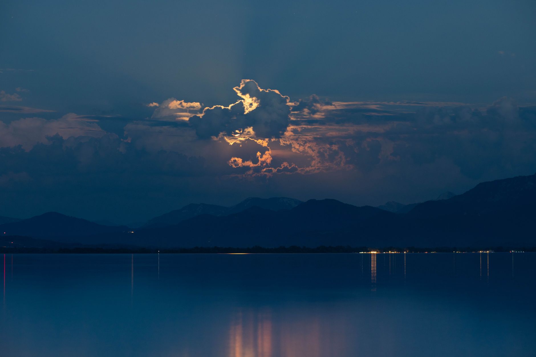 Dramatic night sky with moonlit clouds reflected in a tranquil lake with mountain backdrop.